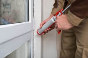 Hands using a caulk gun to apply sealant on cracks near a window sill